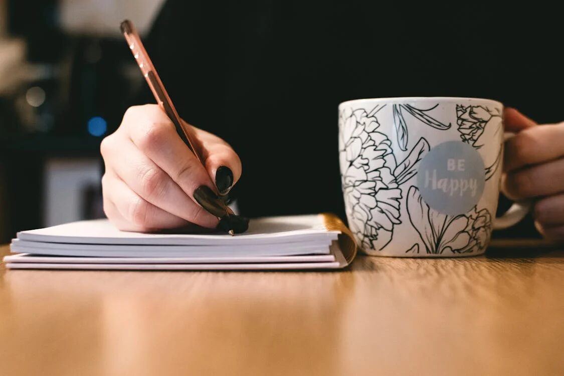 someone writing in a journal holding a cup that says Be happy representing the practice of journaling